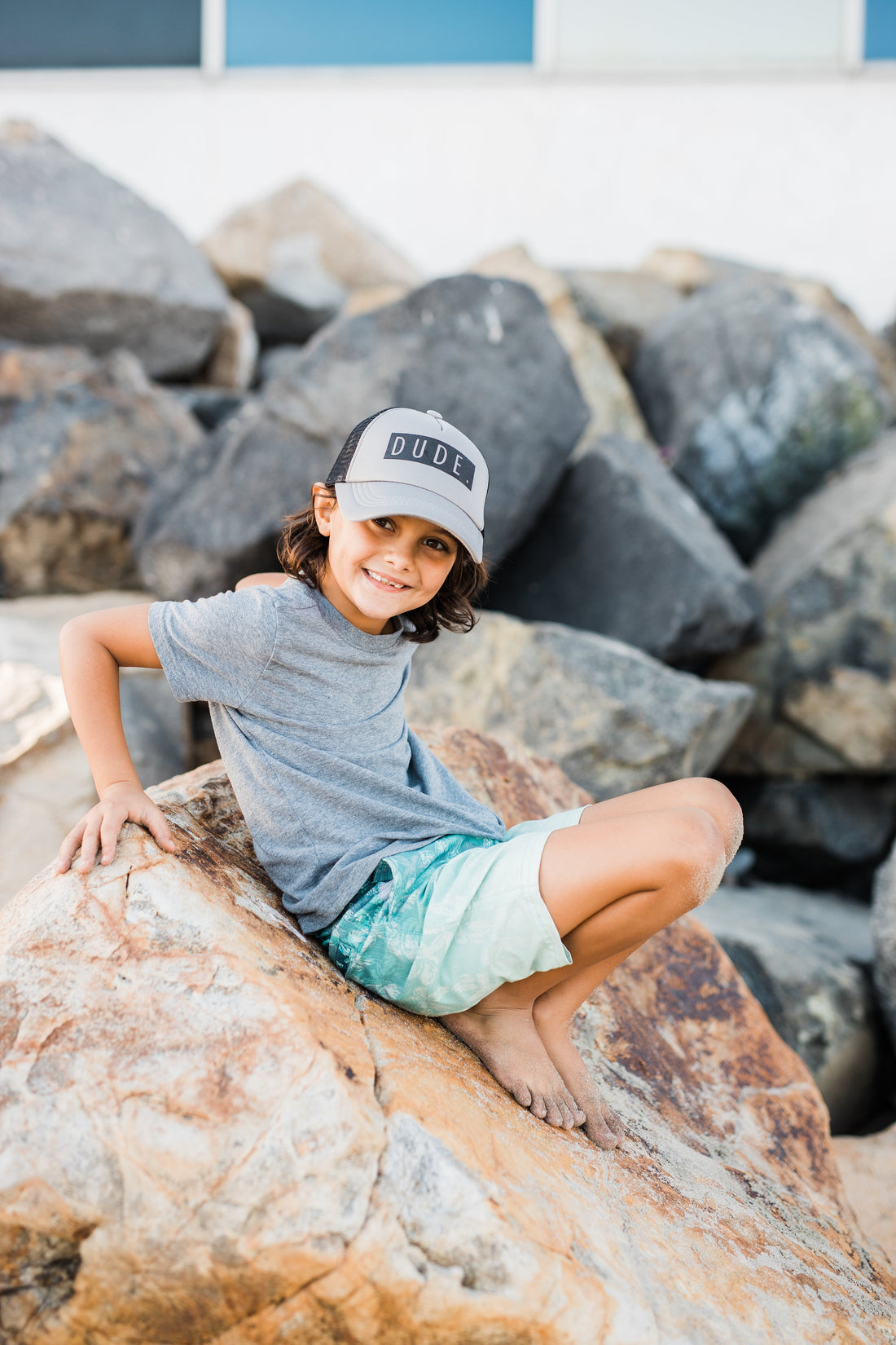 A child wearing a grey and black "DUDE" trucker hat by Mama X™ Brand, along with a grey t-shirt and green shorts, sits barefoot on a large rock outdoors. They are smiling at the camera with large rocks in the background.