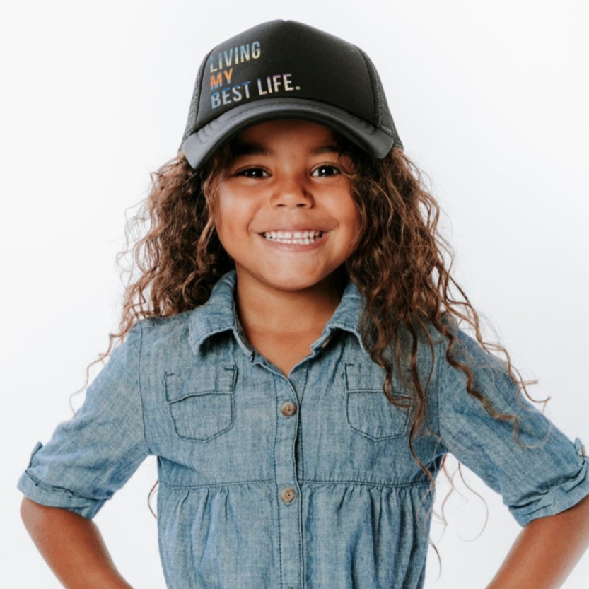 A smiling child with long curly hair wears the "Living My Best Life Trucker Hat" by Mama X™ Brand and a denim button-up shirt. The child is standing against a plain white background, with their hands on their hips.