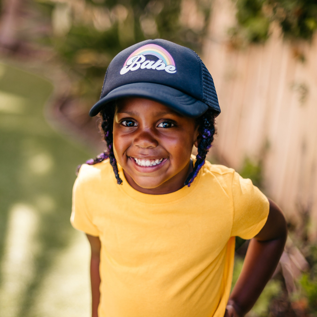 A child with braided hair, wearing a navy blue Rainbow Babe Trucker Hat from Mama X™ Brand, which features a rainbow and the word 