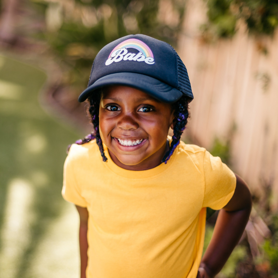 A child with braided hair, wearing a navy blue Rainbow Babe Trucker Hat from Mama X™ Brand, which features a rainbow and the word "Babe", smiles brightly at the camera. The child is dressed in a yellow t-shirt and stands outdoors with greenery and a wooden fence in the background, enjoying sun protection.