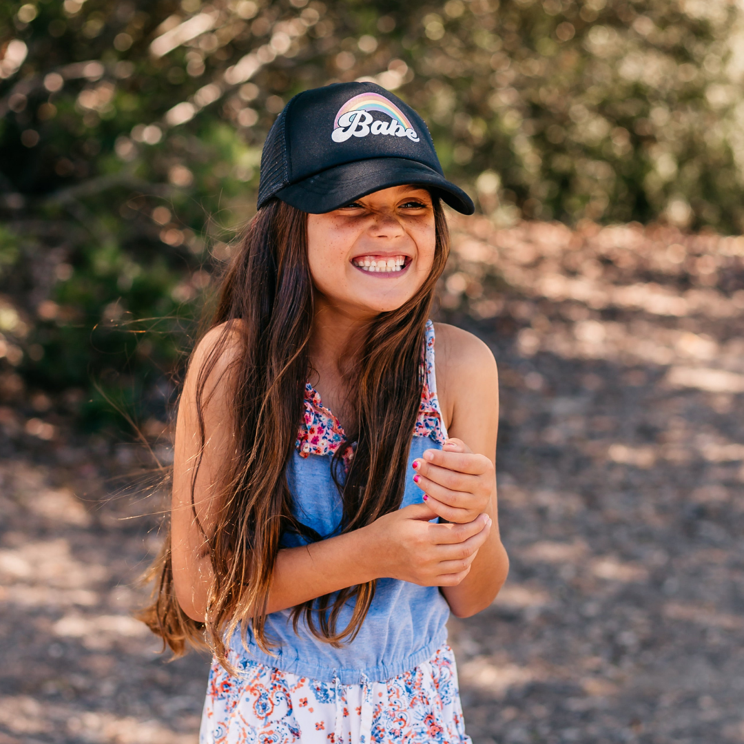 A young girl with long brown hair stands outdoors, smiling widely. She is wearing a colorful floral and denim dress, paired with a Rainbow Babe Trucker Hat by Mama X™ Brand for sun protection. The background is blurred greenery and dirt ground.