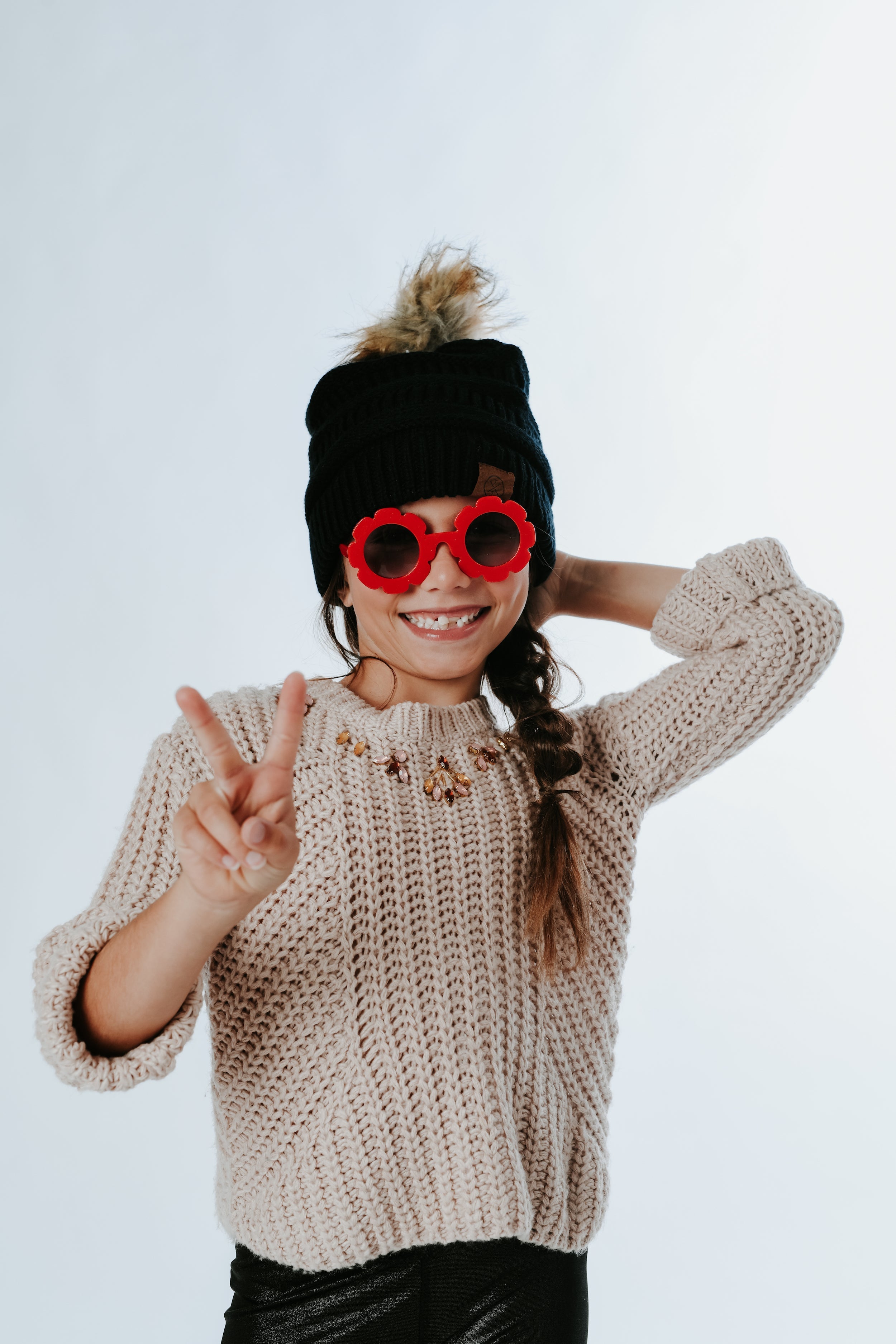 A girl sporting a Black Child Pom Beanie from Mama X Brand, along with a knitted sweater and red flower-shaped sunglasses, smiles playfully while holding up a peace sign.