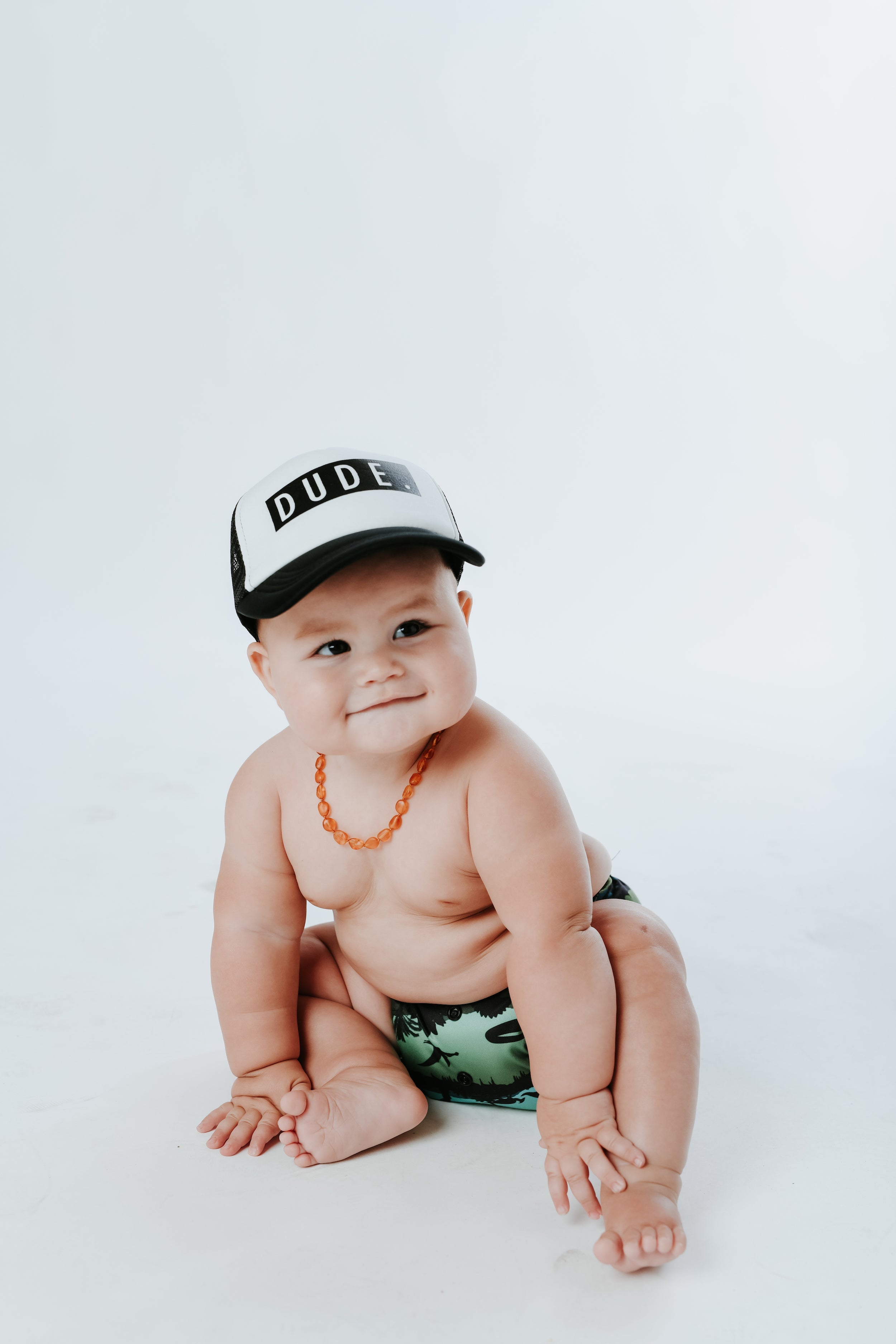 A smiling baby sits on a white background. The baby is wearing a Mama X™ Brand Dude Trucker Hat, an amber-colored necklace, and a green diaper with a leafy pattern. The baby's hands rest on the floor, and they appear content and curious.