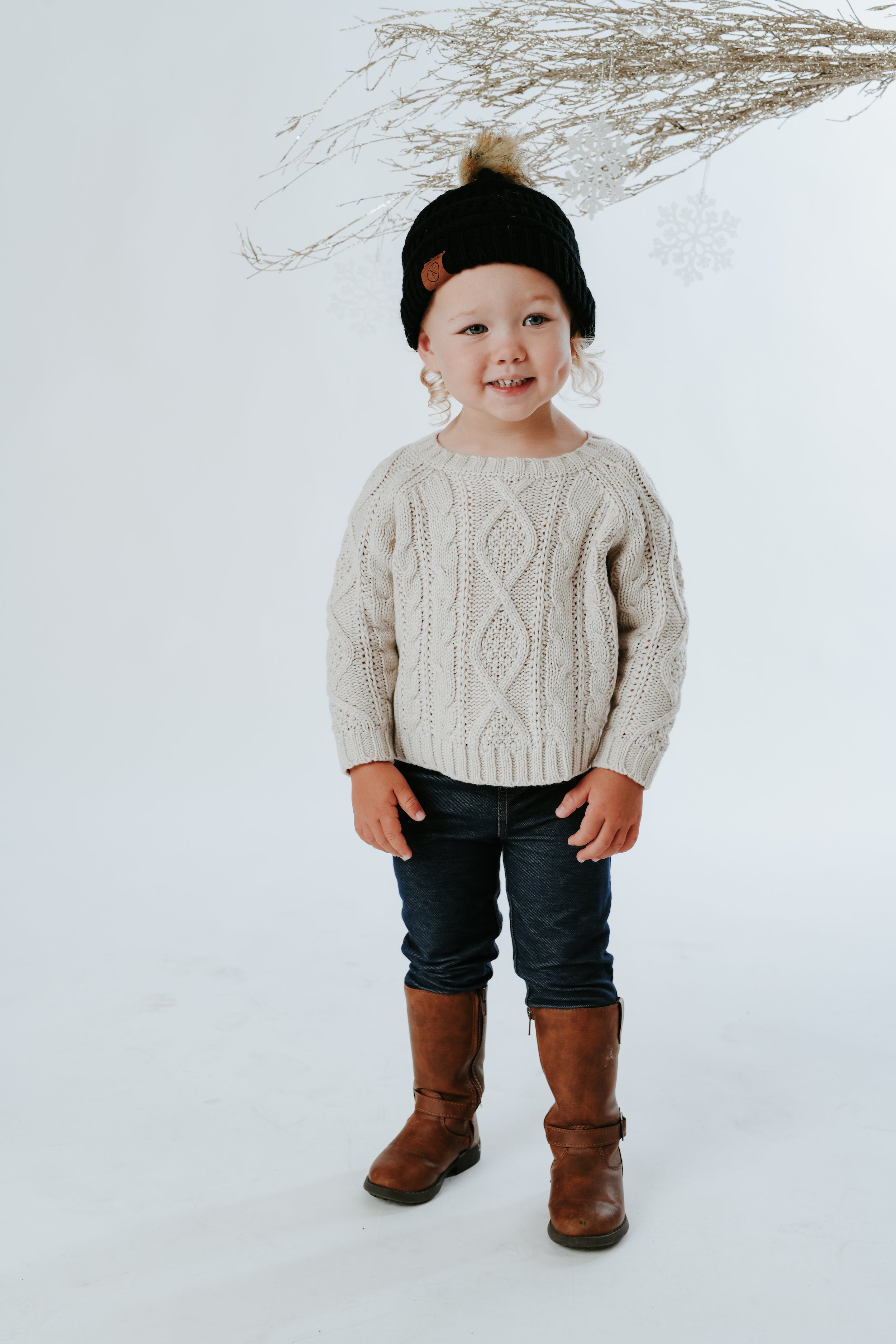 A young child wearing a Mama X Brand Child Pom Beanie in black, accompanied by a cream cable-knit sweater and jeans, stands smiling. They adorn brown boots and are positioned against a light background with dried branches overhead.