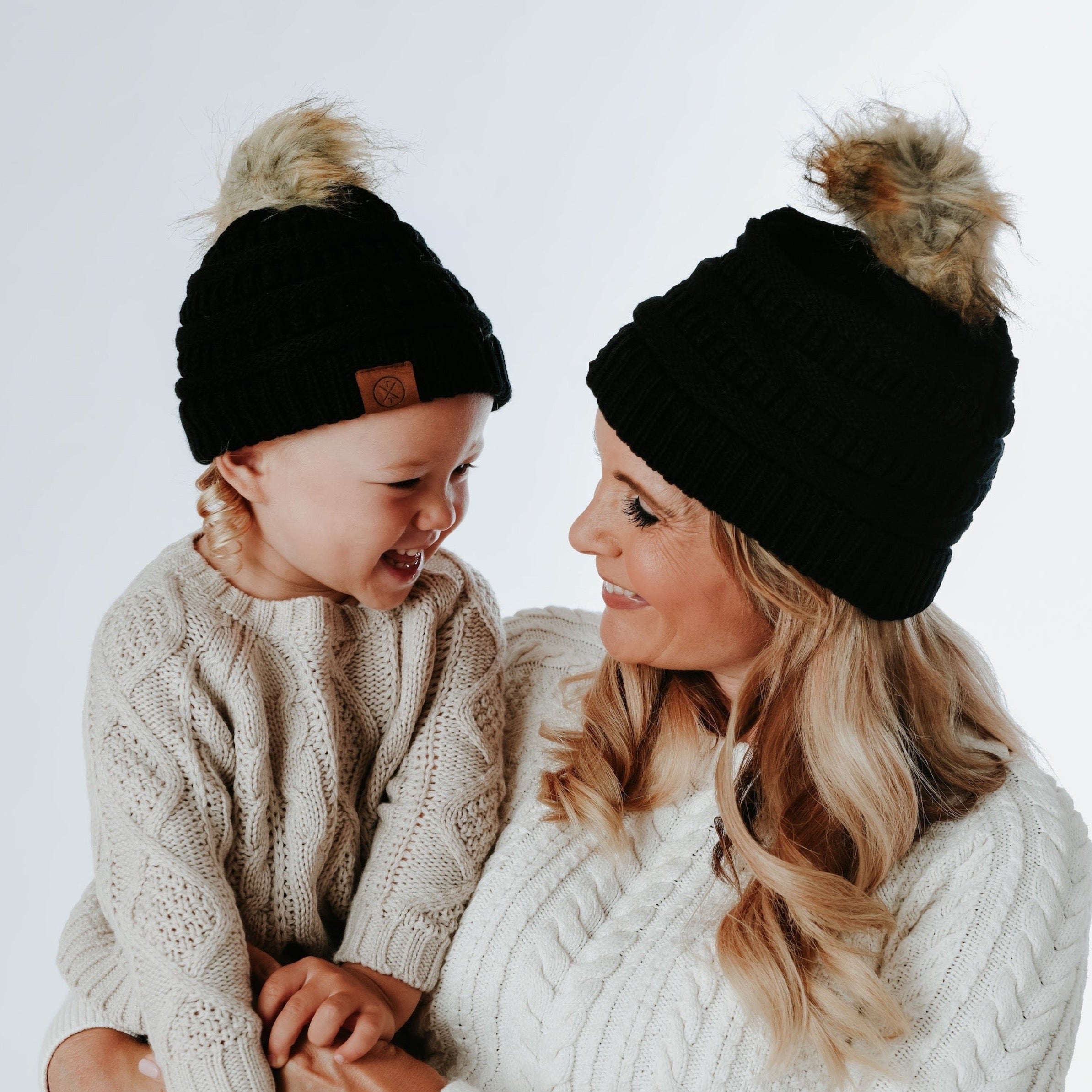 A mother and child, both wearing the Mama X Brand's matching Child Pom Beanie in black, smile warmly at each other. They are dressed in cream-colored cable knit sweaters against a plain background.