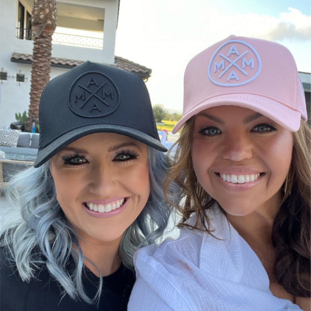 Two women wearing matching Mama X™ Blackout Premium Trucker Hats from the Mama X™ Brand pose for a selfie outdoors. The woman on the left sports a black cap and has wavy blue-gray hair, while the woman on the right wears a pink cap with brown hair. Both are smiling brightly, showcasing their easy maintenance hats.