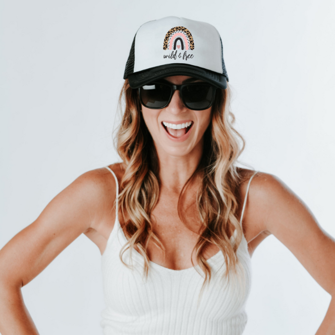 A woman with long, wavy hair smiles broadly. She is wearing sunglasses, a Wild & Free Trucker Hat by Mama X™ Brand featuring an animal print and rainbow design, and a white tank top. Her hands rest on her hips as she poses against a white background.