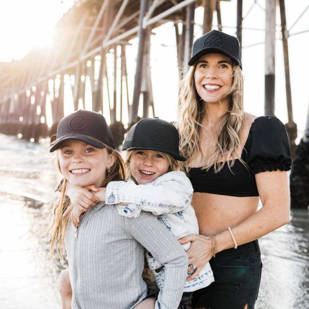 A woman in a black crop top and cap from the Mama X™ Brand's Bro X Blackout Premium Trucker Hat - Stitched 3D Logo collection stands on the beach smiling with her two young children, all wearing matching family hats. One child, in a grey long-sleeve top, smiles while giving a piggyback ride to the other child, dressed in a denim jacket. A pier extends behind them.