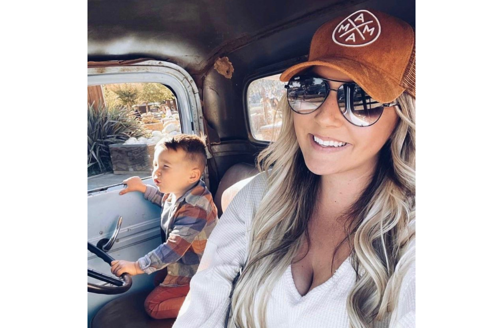 A woman with long blonde hair, wearing a Mama X™ Brand Brown Suede Trucker Hat featuring the embroidered Mama X™ Logo, smiles at the camera. She is seated in a vehicle next to a young boy who is holding the steering wheel and gazing out the window, with outdoor scenery visible in the background.