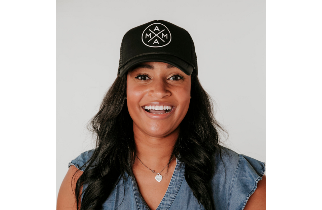 A smiling woman wearing a Mama X™ Foam Trucker Hat, a best-seller from the Mama X™ Brand, and a sleeveless denim top. She has long, wavy dark hair and is adorned with a silver necklace featuring a pendant. The background is plain white.