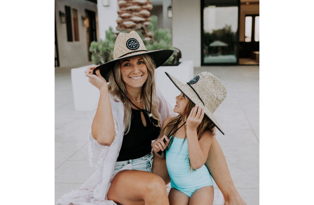 A smiling woman and a young girl sit on the ground outside, both wearing matching Mama X™ Lifeguard Hats for sun protection. The woman is dressed casually in a black top and shorts, while the girl wears a teal swimsuit. They appear happy and playful as they enjoy their time together.