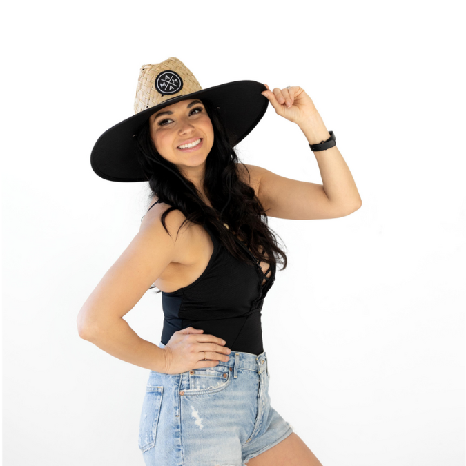 A woman smiles while posing with one hand on her hip and the other holding the brim of her Mama X™ Lifeguard Hat from Mama X™ Brand, offering full-coverage sun protection. She is wearing a black sleeveless top and denim shorts against a plain white backdrop.