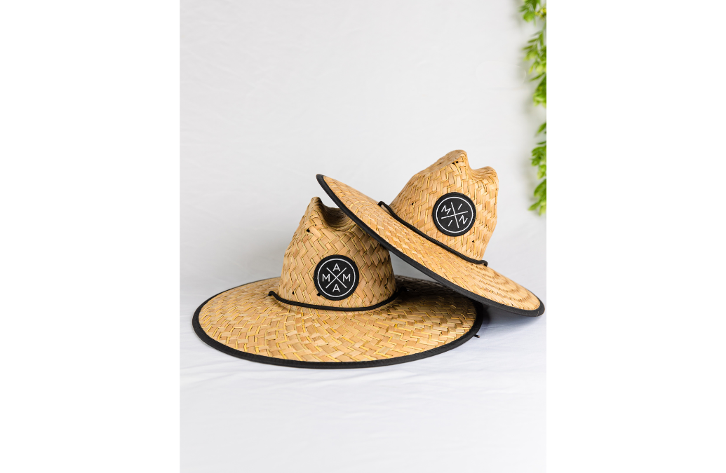 Two Mama X™ Lifeguard Hats from the Mama X™ Brand, featuring wide brims for full-coverage sun protection, are stacked on top of each other against a white background. Each hat displays a black circular patch with a white geometric logo on the front. A green leafy plant is partially visible in the top right corner.