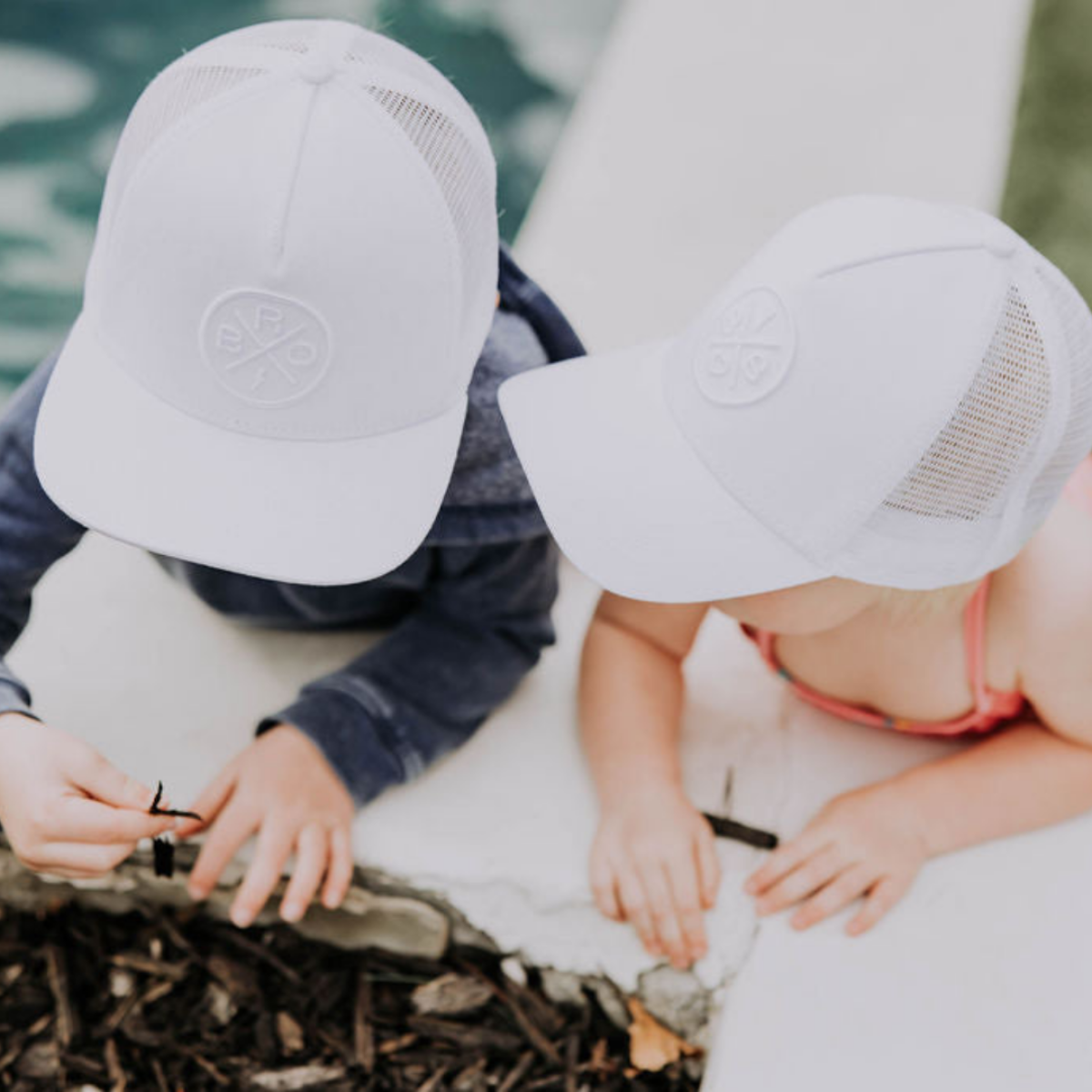 Two children are playing outside, both wearing matching Bro X Whiteout Premium Trucker Hats with a stitched 3D logo from the Mama X™ Brand. They appear to be near a pool, engaged in picking up brown twigs or small objects from the ground. One child is dressed in a light blue shirt, and the other is in a pink outfit.