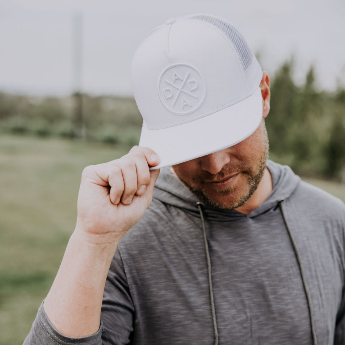 A person wearing a gray hoodie and a Dada X™ Whiteout Premium Trucker Hat with a stitched 3D logo from Mama X™ Brand. The person is tilting their head slightly downwards, touching the brim of the versatile hat, with a blurred outdoor background featuring trees and greenery.