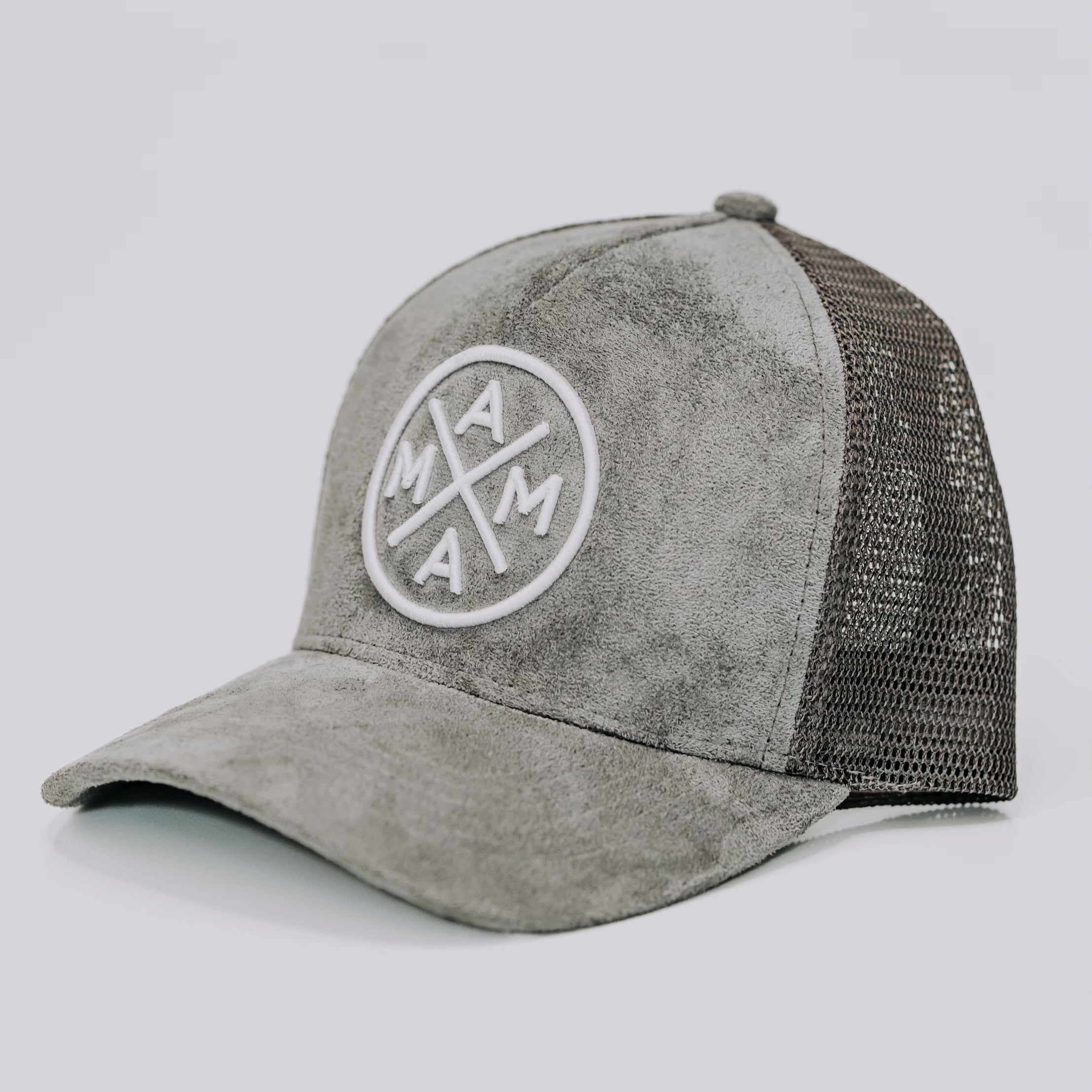 The Mama X™ Trucker Hat - Gray Suede by Mama X™ Brand features a gray suede-like front and a mesh back. The front panel displays a circular emblem with the text "MAMA" in a cross pattern. This stylish 5-panel cap includes an adjustable strap at the back for your convenience.