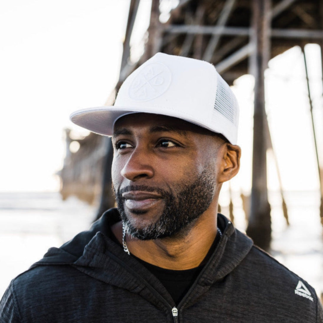 A man with a short beard, wearing a Dada X™ Whiteout Premium Trucker Hat by Mama X™ Brand and a dark hooded jacket, looks to the side. He stands outdoors, likely near a beach or pier, with part of a large wooden structure visible in the background.
