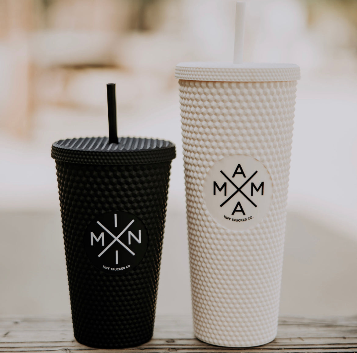 Two textured drink tumblers, one black with a black reusable straw and one larger white with a white straw, sit side by side on a wooden surface. Both BPA-free tumblers feature a circular logo with an 