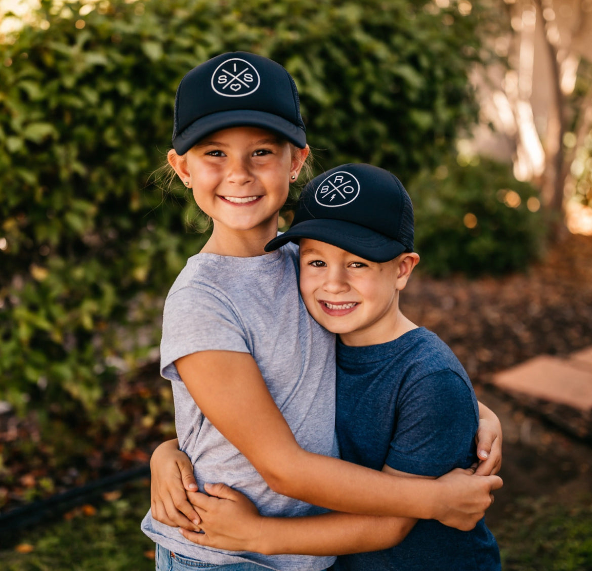 Two children, an older girl and a younger boy, wearing black Bro X Trucker Hats by Mama X™ Brand and casual clothes, embrace each other happily outdoors. They are standing in front of lush greenery and smiling at the camera, enjoying the sun protection their hats provide.