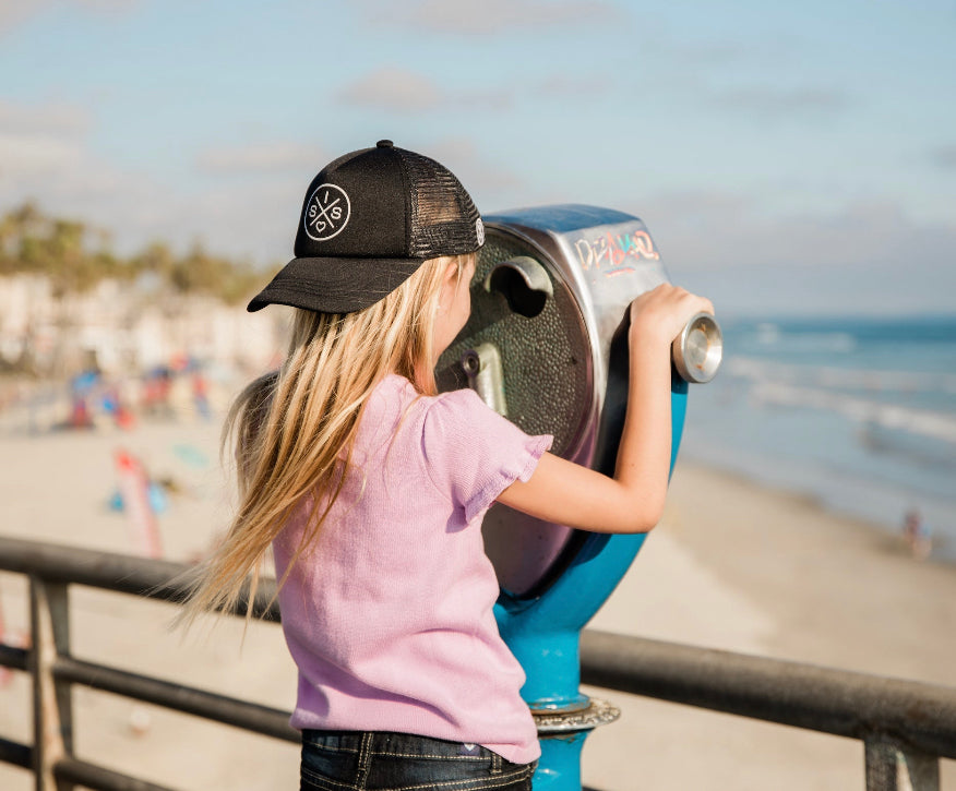 A young girl with long blonde hair, wearing a black Mama X™ Brand Sis X Trucker Hat and a light pink shirt, looks through a blue coin-operated binocular viewer on a pier, overlooking a sandy beach and ocean on a sunny day.