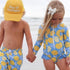 Two young children holding hands at the beach. The child on the left is wearing a "Ray of Sunshine Trucker Hat" from Mama X™ Brand, which provides excellent sun protection, and a blue swimsuit with yellow lemon prints. The child on the right is wearing a matching blue and yellow lemon print swimsuit.