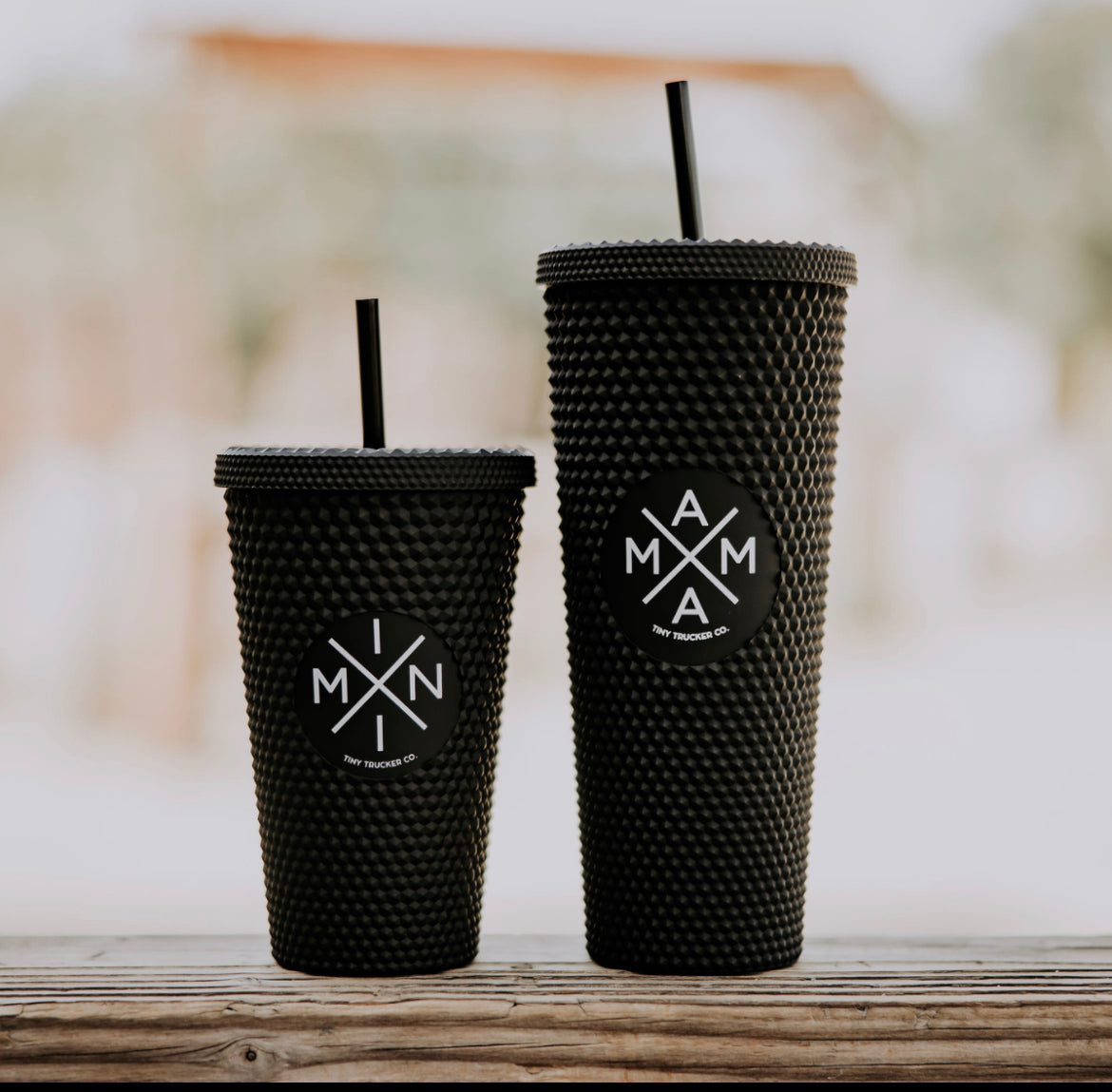 Two black, textured travel tumblers with reusable straws stand side by side on a wooden surface. The smaller tumbler, the "Mini X Studded Plastic Tumbler 16oz" from Mama X™ Brand, and the larger one labeled "MAMA," both boast a BPA-free, modern minimalist design against a blurred outdoor background.