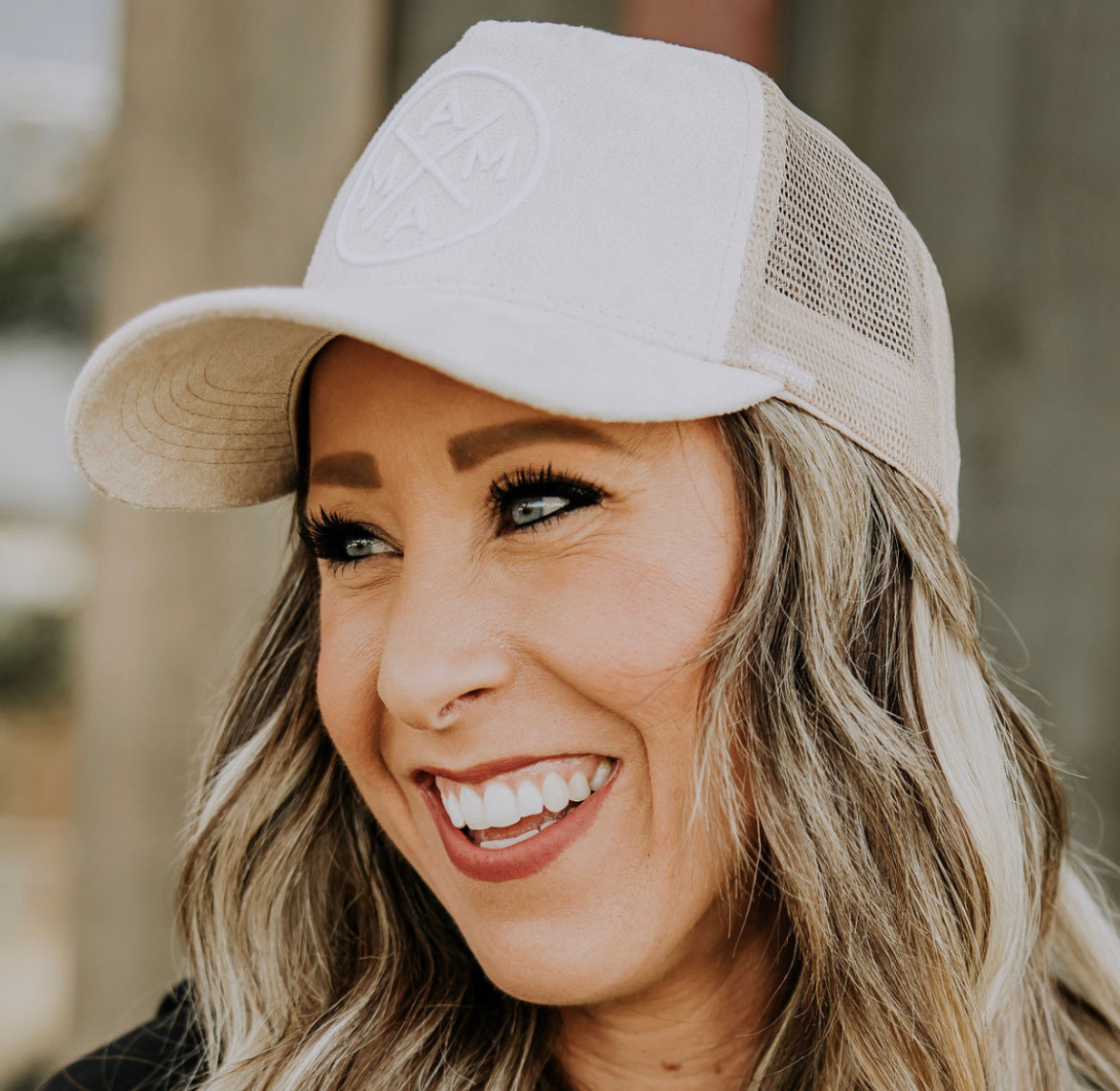 Close-up of a person smiling, wearing a beige Mama X™ Light Suede Trucker Hat with a circular logo and mesh back. The person has long, wavy hair and is outdoors, with a blurred background.