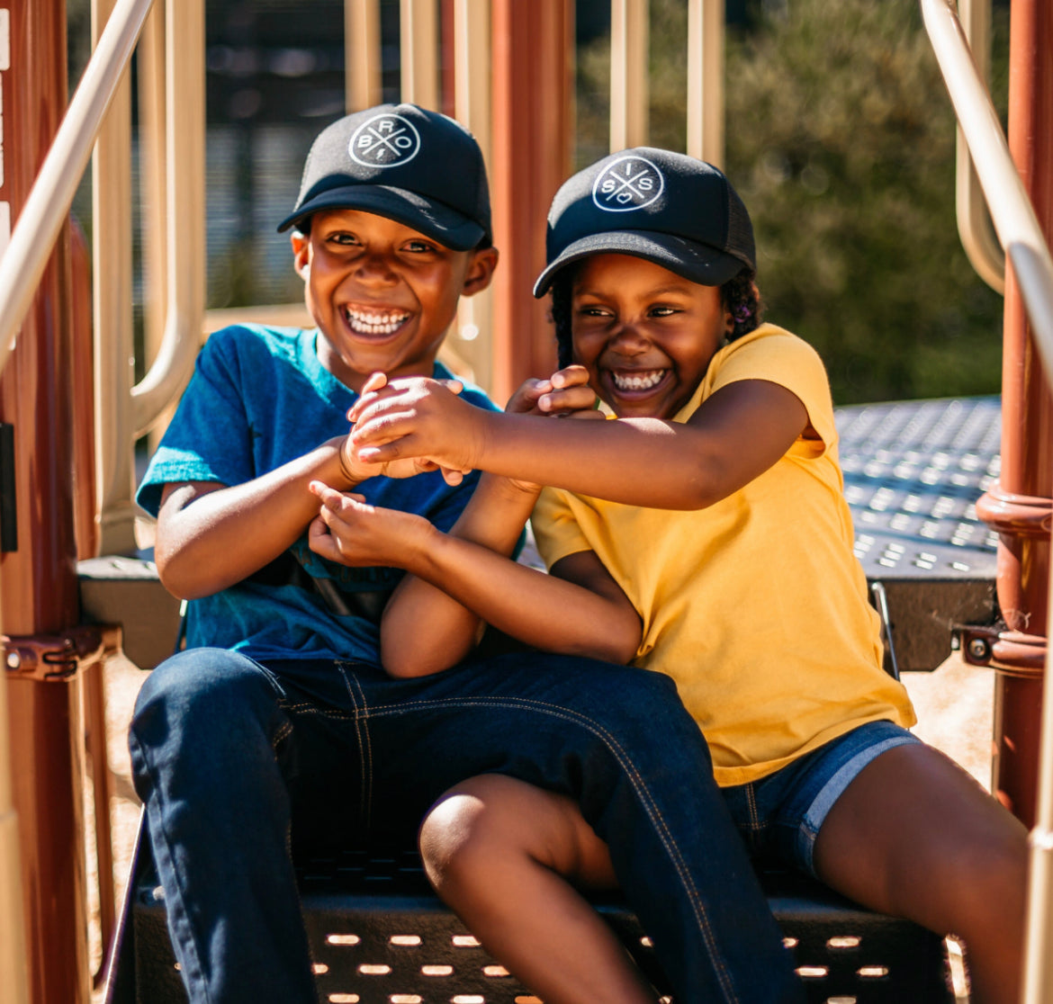 Two children sit on playground steps, smiling and holding hands. One child wears a blue shirt and jeans, while the other sports a yellow shirt and denim shorts. Both are wearing Bro X Trucker Hats from Mama X™ Brand, featuring a white circular logo that offers stylish sun protection. The background is blurred, highlighting play equipment.