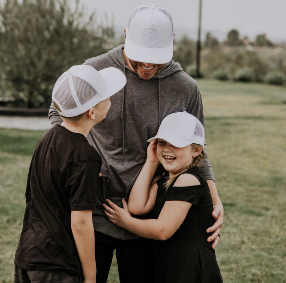 A man wearing a gray hoodie and a Mama X™ Brand Bro X Whiteout Premium Trucker Hat - Stitched 3D Logo stands outdoors with two smiling children. The boy, also in a white cap and black shirt, looks up at the man. The girl in a white cap and black dress laughs while hugging the man. All are enjoying the grassy area under the sun&