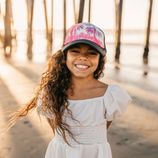 A young girl with long curly hair smiles at the camera while standing on a beach at sunset. She is wearing a white dress and a vibrant Sunny Palms Trucker Hat by Mama X™ Brand for sun protection. The wooden pillars of a pier are visible in the background.