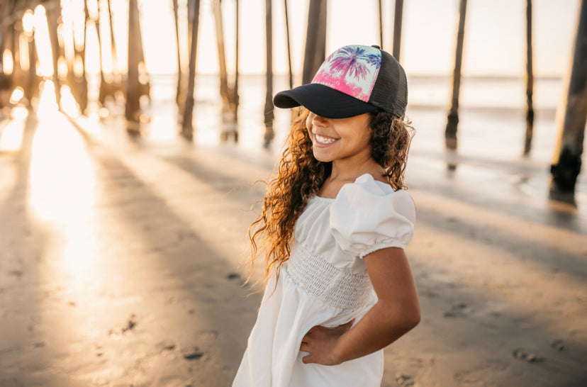 A young girl with long curly hair smiles while standing on a beach. She wears a white dress and a Sunny Palms Trucker Hat from Mama X™ Brand, featuring a tropical design with pink and blue palm trees and providing her with stylish sun protection. The background shows the sun setting through large wooden pillars of a pier.