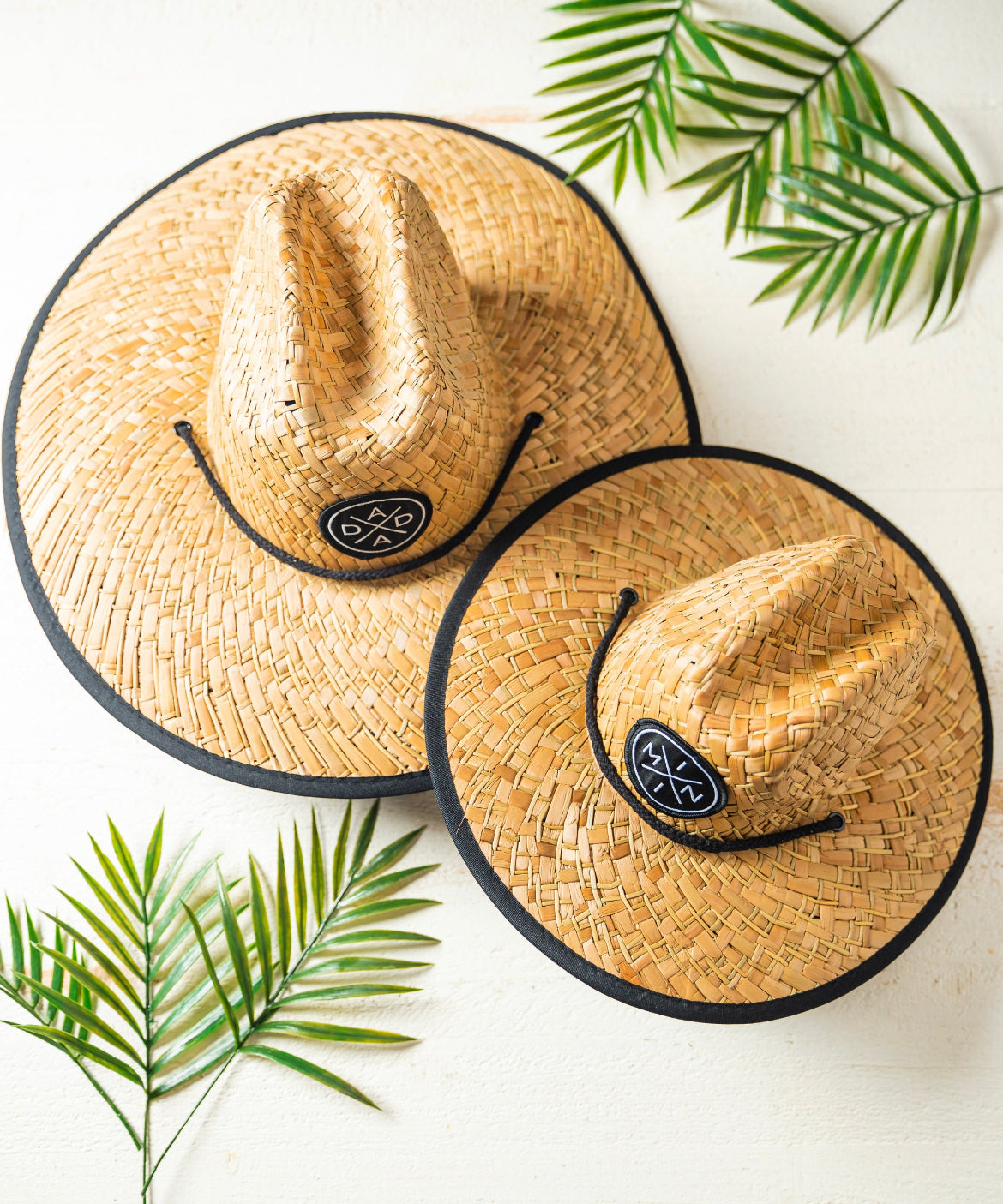 Two Dada X™ Lifeguard Hats by Mama X™ Brand, each featuring black circular patches with a white geometric pattern, are placed on a light-colored surface. The hats have black edges and provide excellent sun protection. Several green tropical leaves add a fresh and summery vibe to the scene.