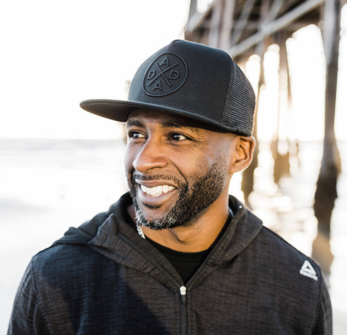 A man wearing a Mama X™ Brand Dada X™ Blackout Premium Trucker Hat with a stitched 3D logo and a dark jacket stands near a wooden pier. He has a short beard and is smiling while looking off to the side. The background includes a beach scene with the ocean and part of the pier structure.