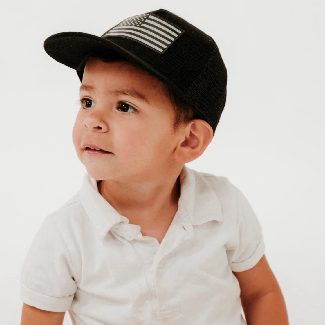 A young child wearing a white polo shirt and a Mama X™ Brand American Flag Leather Patch Trucker Hat looks to the side against a plain white background, with a thoughtful expression on their face.
