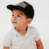 A young child wearing a white polo shirt and a Mama X™ Brand American Flag Leather Patch Trucker Hat looks to the side against a plain white background, with a thoughtful expression on their face.