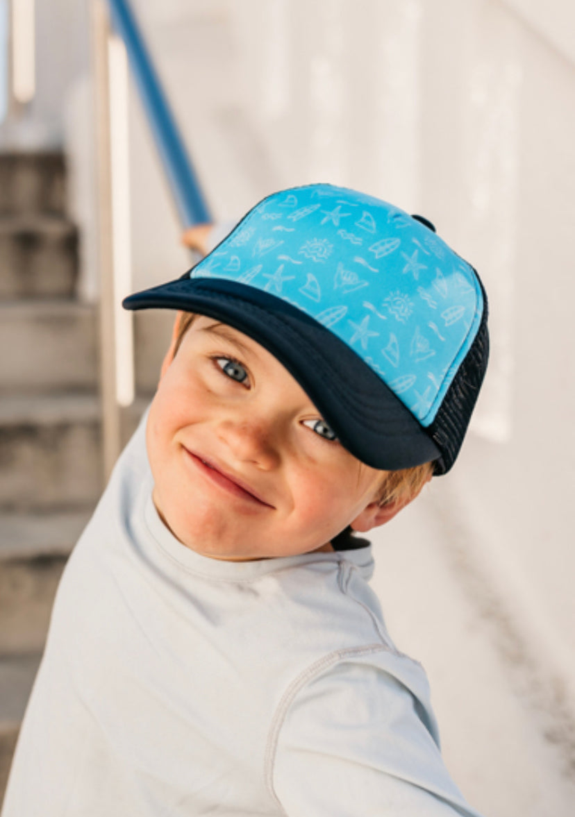 A cheerful young boy, wearing a Shaka Repeater Trucker Hat from Mama X™ Brand and a white shirt, is smiling at the camera. He stands in a sunny outdoor setting, with steps and a railing visible in the background.