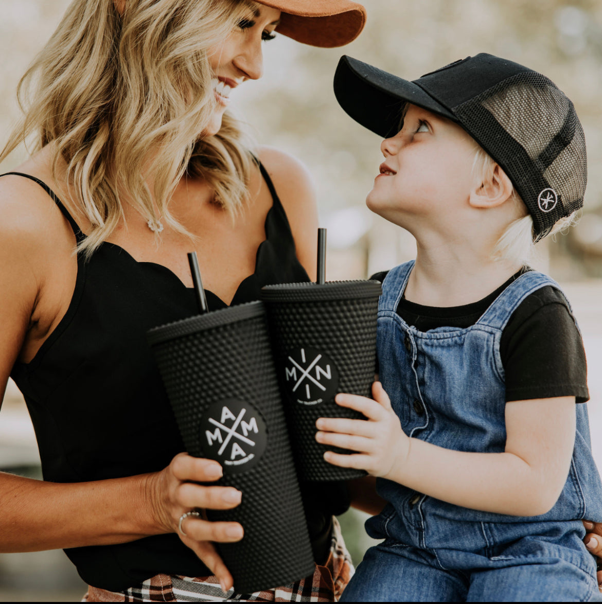 A woman and a child, both wearing hats, smile at each other while holding black Mini X Studded Plastic Tumblers, 16oz from the Mama X™ Brand, featuring reusable BPA-free straws and a round logo. The woman is dressed in a black tank top, and the child wears a black shirt with denim overalls. The background is blurred, suggesting an outdoor setting.