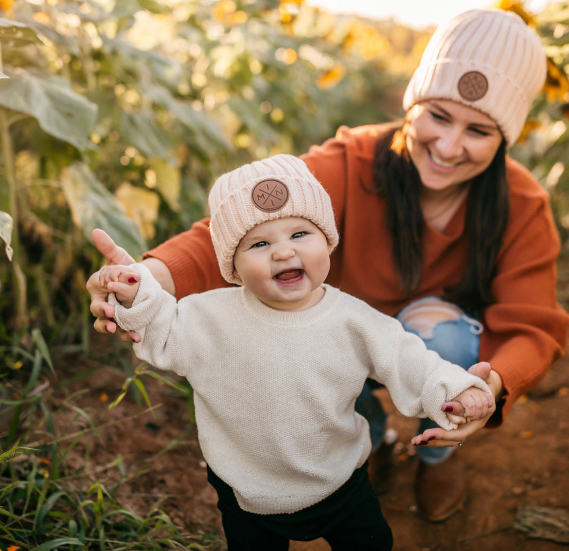 A smiling baby wearing a Mama X™ Brand beige Knit Beanie With Brown Leather Patch and a white sweater holds hands with an equally joyful adult, who is also wearing a matching Mama X™ Brand knit beanie along with an orange sweater, in the midst of a sunflower field. The adult is kneeling behind the baby, offering support.