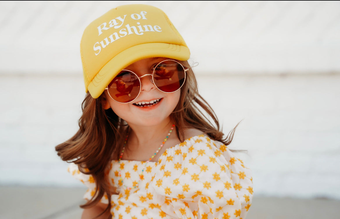 A young girl wears a "Ray of Sunshine" Trucker Hat by Mama X™ Brand, along with round sunglasses and a yellow floral-patterned dress. She smiles brightly while standing outdoors, embracing the sun&