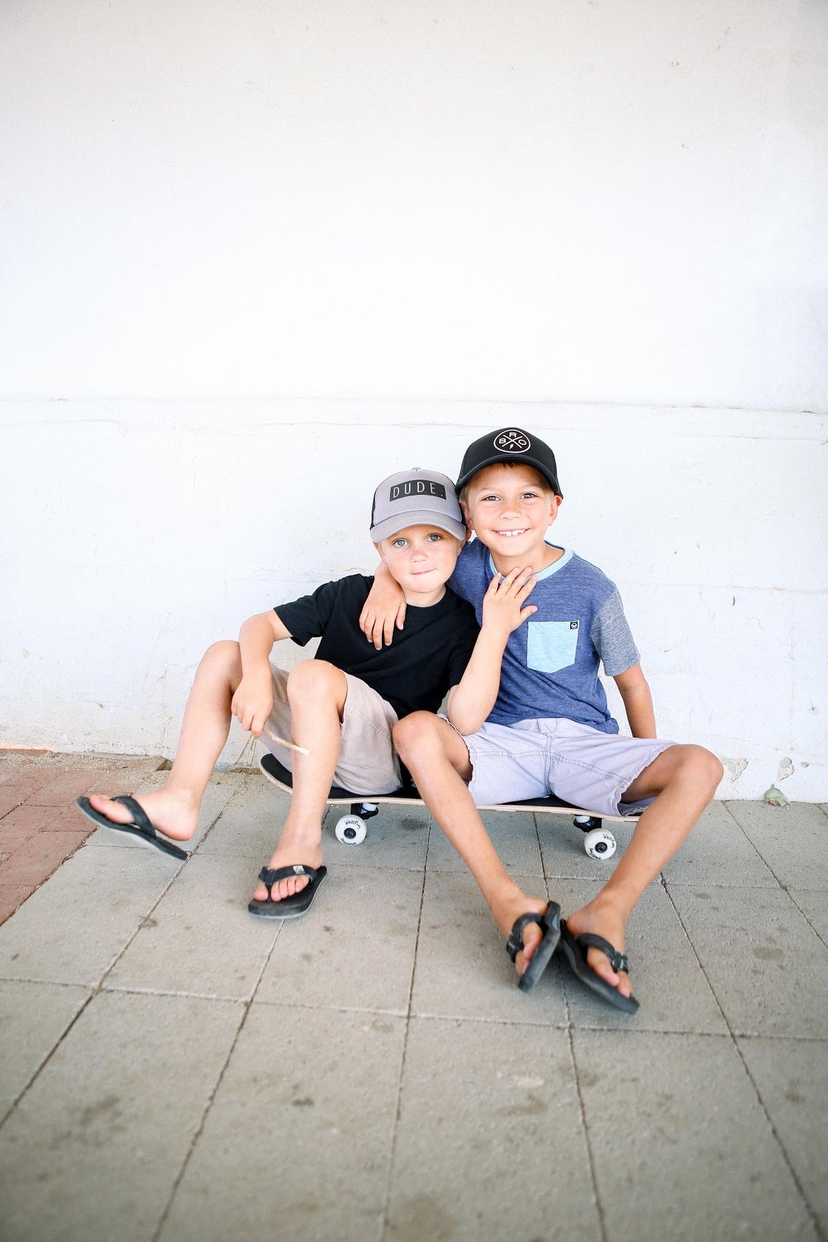 Two boys are sitting on a skateboard against a white wall. The boy on the left is wearing a black t-shirt, khaki shorts, an adjustable snap cap, and flip-flops. The boy on the right is in a blue t-shirt, light blue shorts, a 5-panel cap, and flip-flops. They are both smiling. The snap cap worn by the boy on the left appears similar to the DUDE Trucker Hat - Grey & Black from Mama X™ Brand.