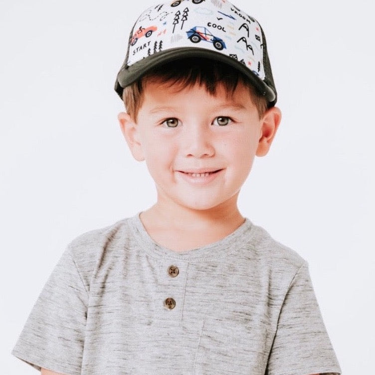 A young child with short brown hair smiles at the camera, wearing a light gray shirt with button details and a Mama X™ Brand Cars Trucker Hat, which is adorned with colorful drawings of cars and the word "COOL." The background is a plain, light color.