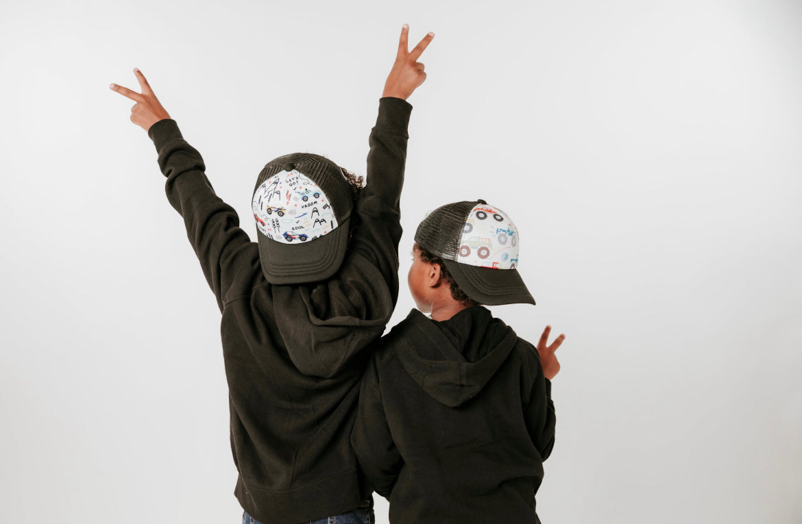 Two children in black hoodies and vibrant Mama X™ Brand Cars Trucker Hats stand against a plain background with their backs facing the camera. Both raise their hands, making peace signs towards the sky.