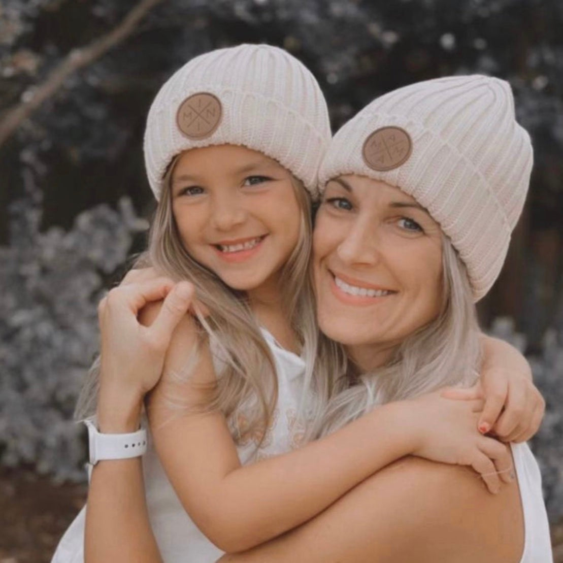 A smiling woman and a young girl, possibly her daughter, are wearing matching Mama X™ Brand Knit Beanies with a brown leather patch. Hugging the girl and looking at the camera, they appear to be outdoors with foliage blurred in the background. Their cozy attire is a perfect Fall and Winter staple.