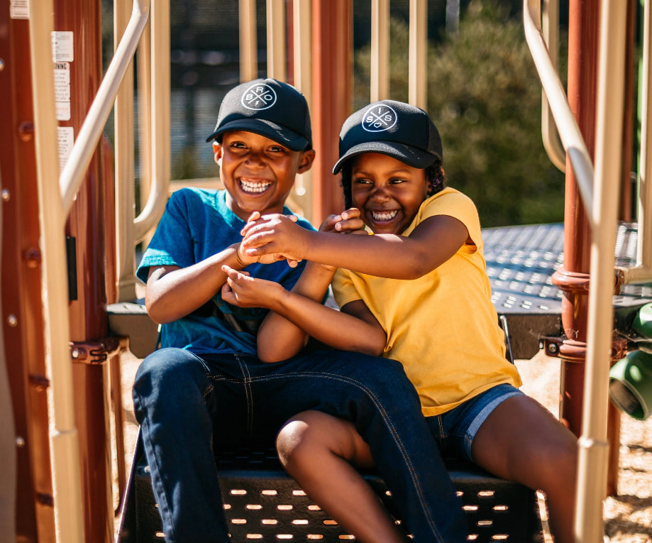 Two children, a boy and a girl, are sitting on a playground structure. Both are smiling and wearing black Sis X Trucker Hats by Mama X™ Brand. The boy is in a blue shirt and jeans, while the girl sports a yellow shirt and shorts. Their 5-panel caps add a trendy touch as they happily enjoy their playtime together.